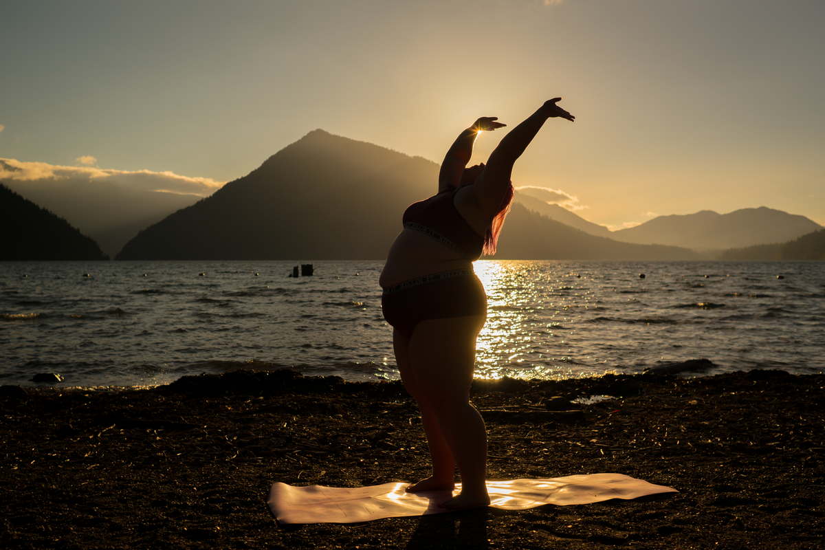 A fat woman with pink hair stands on a yoga mat and throws her arms joyfully into the sky as she begins to move into a yoga pose. A beautiful lake shore, distant mountains and sunset glow complete the image.