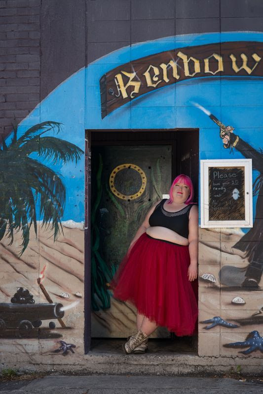 A plus-size femme person with bright pink hair, a tutu skirt and crop top leans against the doorway of a building painted in a pirate theme.
