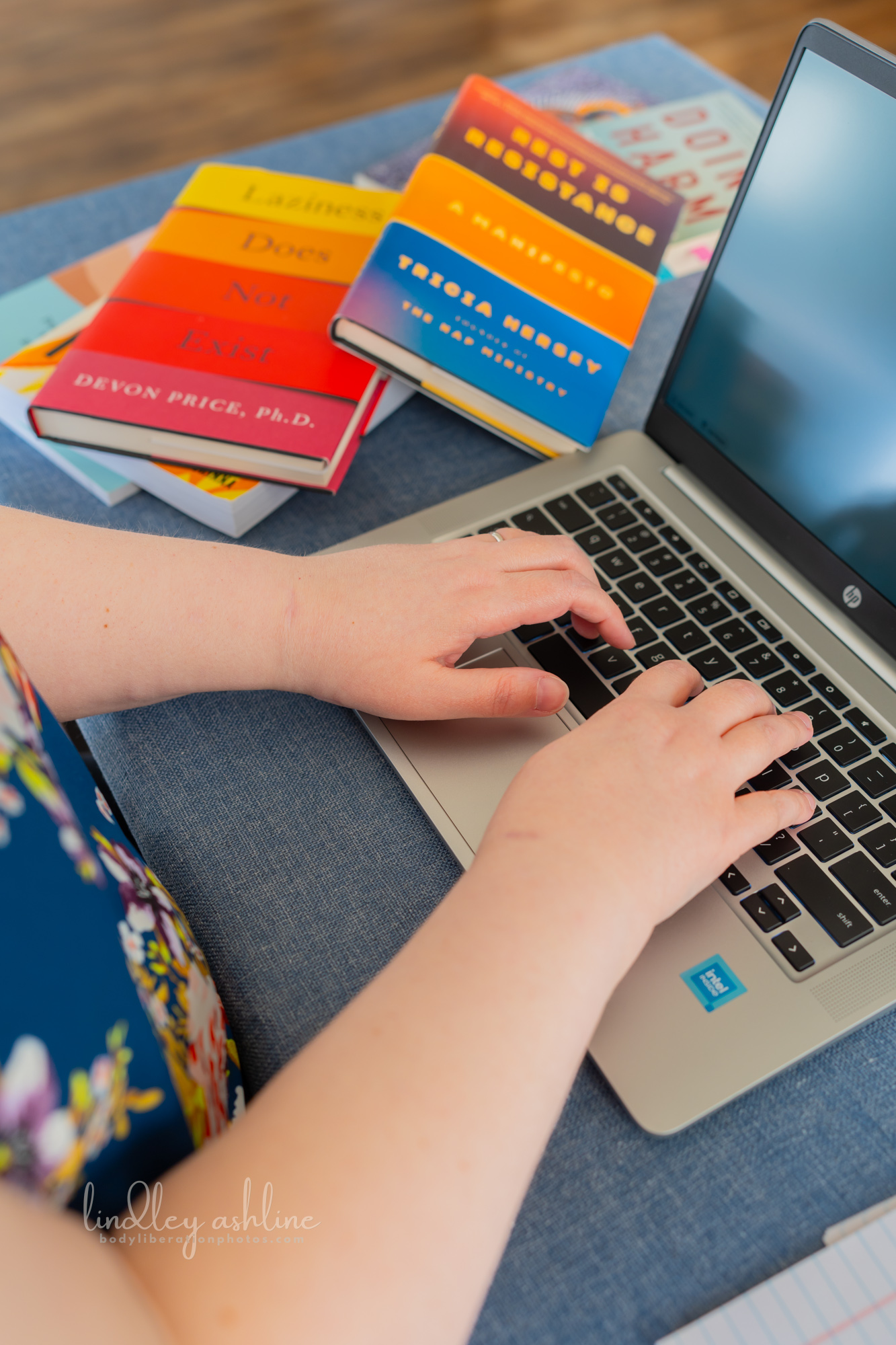 A plus-size white woman types during a telehealth therapy session in Washington state at a Pacific Northwest fat-positive professional portrait photo shoot. Copies of the books Laziness Does Not Exist and Rest is Resistance can be seen behind her.
